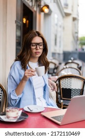 Stylish Woman Holding Cup Near Laptop And Blurred Dessert On Cafe Terrace In Paris