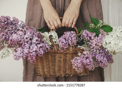 Stylish woman holding wicker basket with beautiful lilac flowers in rustic room. Female in linen dress arranging lilac flowers, cropped view. Authentic moody moment. Rustic wedding - Powered by Shutterstock