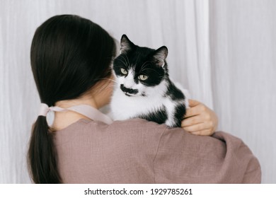Stylish woman in linen dress holding cute cat on background of pastel fabric. Simple slow living. Young female in boho rustic dress hugging adorable black and white kitty, lovely moment