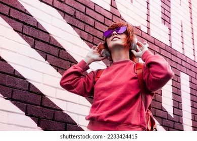 Stylish woman in magenta color jacket and heart shaped sunglasses wearing wireless headphones on her head and listening music. Urban city street fashion. Color of the 2023 year. Selective focus. - Shutterstock ID 2233424415