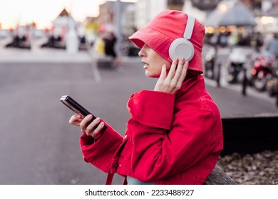 Stylish woman in magenta color jacket and bucket hat wearing wireless headphones on her head and setting phone for listening music. Urban city street fashion. Color of the 2023 year. Selective focus - Shutterstock ID 2233488927
