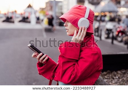Stylish woman in magenta color jacket and bucket hat wearing wireless headphones on her head and setting phone for listening music. Urban city street fashion. Color of the 2023 year. Selective focus