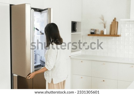 Stylish woman opening fridge and looking inside in new minimal white kitchen. Housewife cleaning up kitchen in new modern scandinavian home. Food and diet concept