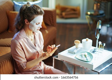 Stylish Woman In Pajamas With White Sheet Facial Mask On Face Cloud Download To Smartphone From Stored Data On Server In The Modern Living Room In Sunny Winter Day.