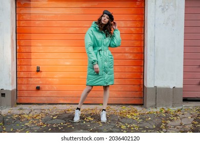 Stylish Woman Posing In Winter Autumn Fashion Trend Puffer Coat And Hat Beret Against Orange Wall In Street Wearing Sneakers