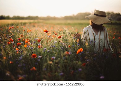 Stylish woman in rustic  dress and hat walking in summer meadow among poppy and wildflowers in sunse