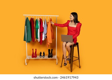 A stylish woman sits on a tall stool, holding a laptop, while thoughtfully examining a selection of colorful autumn clothing hung on a rack, with a range of shoes lined up below - Powered by Shutterstock