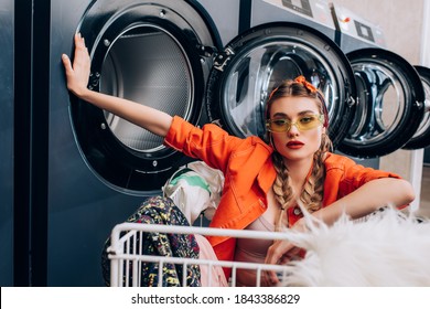 stylish woman sitting in cart with clothing near washing machines in laundromat - Powered by Shutterstock