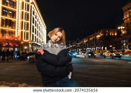 Stylish woman stands on the street and poses for the camera against the backdrop of the night city and street architecture decorated for the holidays.