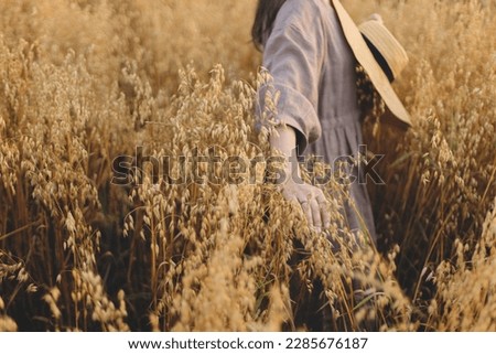 Stylish woman with straw hat holding oat stems in evening light. Rural slow life. Young female in rustic linen dress standing in harvest field in summer countryside. Atmospheric tranquil moment