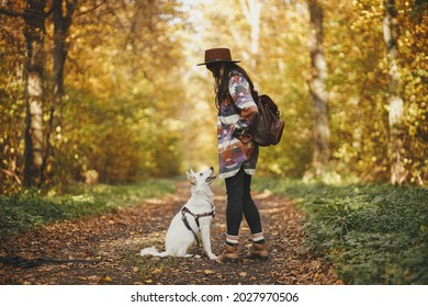 Stylish Woman Traveler In Hat Hiking With Cute Dog In Sunny Autumn Woods. Young Female Hipster Traveling With Swiss Shepherd White Dog. Travel With Pet, Loyal Companion. Wanderlust