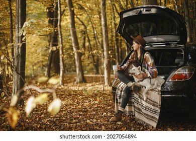 Stylish Woman Traveller With Backpack Sitting With Cute Dog In Car Trunk In Sunny Autumn Woods. Young Hipster Female Traveling With Swiss Shepherd White Dog. Travel And Road Trip With Pet