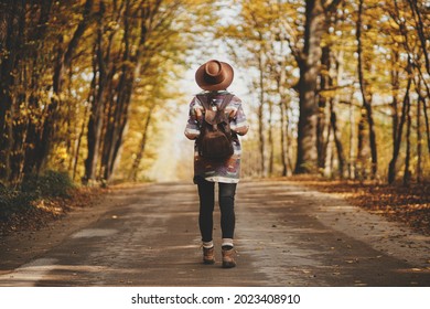 Stylish Woman Traveller In Hat With Backpack Walking On Road In Sunny Autumn Woods. Young Female Hipster Hiking In Fall Forest, Beautiful Moment. Travel And Wanderlust Concept. Back View