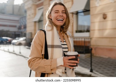 stylish woman walking in winter street wearing beige coat, scarf, smiling happy cold season fashion trend, holding coffee cup - Powered by Shutterstock