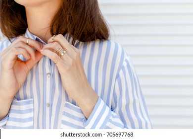 Stylish woman wearing blue striped shirt buttoning a button. Details of trendy casual outfit. Street fashion. Gold rings on female fingers. Jewelry, accessories, close up, women hands.