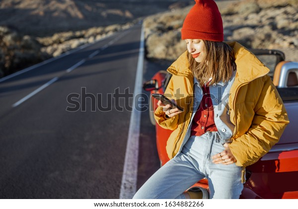 Stylish woman in yellow jacket and red\
hat enjoying roadtrip, sitting with a smart phone on the car hood\
on the roadside of the desert valley during a\
sunset