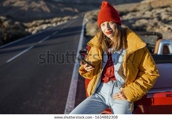 Stylish woman in yellow jacket and red\
hat enjoying roadtrip, sitting with a smart phone on the car hood\
on the roadside of the desert valley during a\
sunset