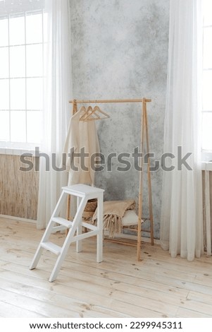 Stylish wooden coat rack and white wooden ladder stool in a spacious interior with large windows with curtains. Nobody. Wooden floor in the apartment