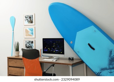 Stylish workplace with modern computer and SUP board near light wall in room. Interior design