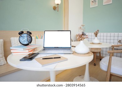 A stylish workspace setup featuring a laptop mockup, alarm clock, books, and stationery on a round table in a pastel green cafe interior, perfect for remote work and productivity concepts.
