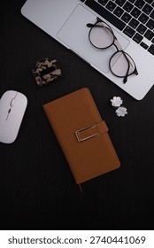 A stylish workspace setup: silver laptop with glasses on the trackpad, a white wireless mouse, a brown leather notebook, a hair clip, and delicate white flowers earings on a dark desk. 