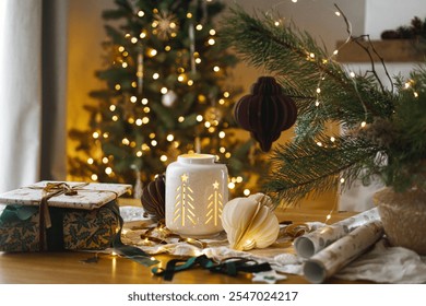 Stylish wrapped christmas gift, glowing lantern and modern paper ornaments on rustic table on background of christmas tree lights bokeh. Merry Christmas! Atmospheric christmas time - Powered by Shutterstock