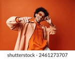 A stylish young African American man with curly hair wearing an orange shirt and sunglasses, posing energetically against an orange background.