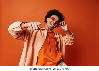 A stylish young African American man with curly hair wearing an orange shirt and sunglasses, posing energetically against an orange background.