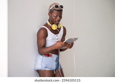 Stylish young african american non-binary person using digital tablet while wearing headphones and sunglasses