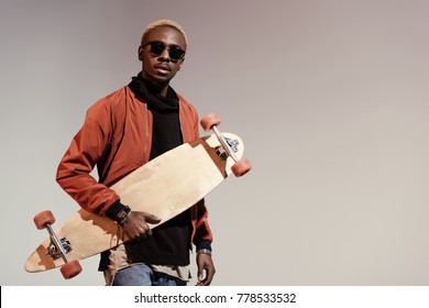 Stylish young african american skater holding longboard in hand isolated on light background