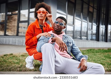 Stylish young african american woman in bright clothes wearing sunglasses and looking away near confident best friend sitting on border on urban street, stylish friends enjoying company - Powered by Shutterstock