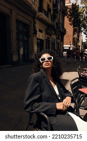 Stylish Young African Girl With Curly Hair Looking At Camera Sitting On Chair In Cafe Outside. Brunette Hair Model Wears Dress And Jacket. Concept Sunshine, Vacation.