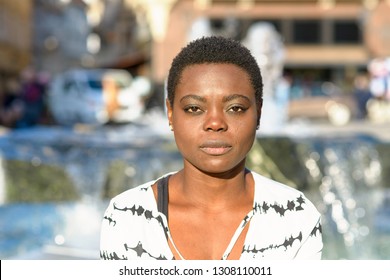 Stylish Young African Woman With A Calm Deadpan Expression Staring Quietly At The Camera Standing In Front Of A City Fountain