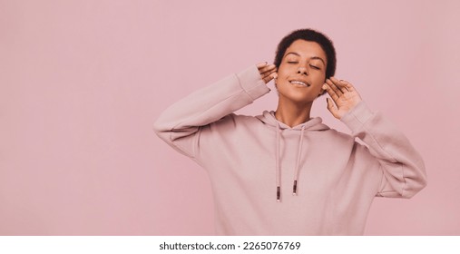 Stylish young african woman in hoodie lstening to music in wireless headphones. Lifestyle concept with copy space. Girl standing with closed eyes and enjoying songs over pink pastel backdrop