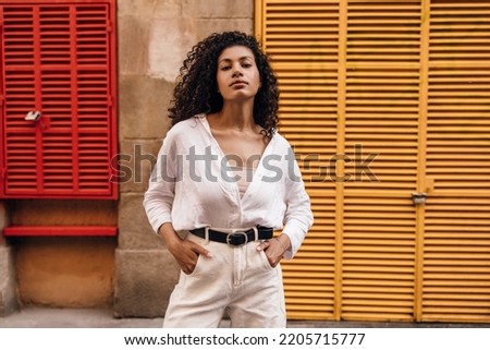 Stylish young african woman looking at camera posing with hands in pockets outdoor. Brunette wears singlet, shirt and white jeans. Concept female beauty.
