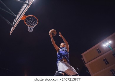 Stylish young basketball player in cap is jumping and shooting a ball through the hoop while playing outdoors at night - Powered by Shutterstock