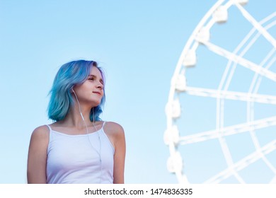 Stylish Young Beautiful Hipster Woman Listening To Music On Her Mobile Phone Using Headphones In An Amusement Park Enjoying A Sunny Day, Cool Accessories. Carousels And Ferris Wheel