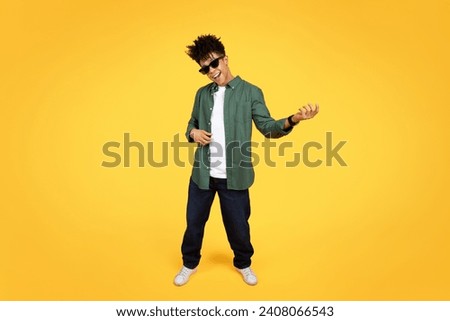 Stylish young black guy wearing sunglasses and casual clothing playing imaginary guitar isolated on yellow studio background. Cool african american millennial man rockstar, copy space, full length