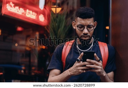 Stylish young Black man using smartphone outside restaurant, focused on screen, casual look with red backpack and jewelry, modern tech lifestyle and mobile finance theme