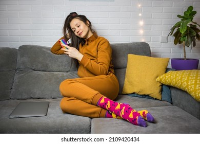 Stylish young brunette woman in warm brown tracksuit, colorful socks is lying relaxed on gray couch with smartphone in hand. A girl is using smartphone, gadget, online shopping, chatting. Cozy home.