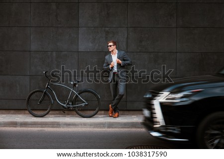 Stylish young businessman in sunglasses stands near a bicycle with his back against a brickgray wall