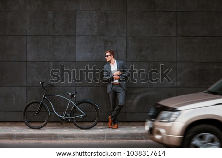 Stylish young businessman in sunglasses stands near a bicycle with his back against a brickgray wall
