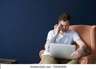 Stylish Young Caucasian Male Freelancer With Beard And Tattoos On Arms Feeling Frustrated Because Of Headache, Can't Focus On Work, Sitting On Comfortable Orange Armchair, Using Laptop Computer.