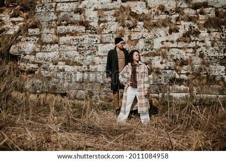 stylish young couple in casual style in the fields in spring against the background of a textured stone wall, they tenderly hug and fool around. Image with selective focus 