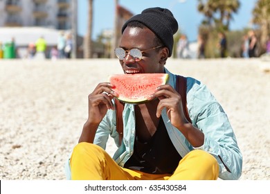 Stylish Young Dark-skinned European Man Refreshing Himself On Summer Day, Sitting Alone On Pebble Beach And Eating Ripe Watermelon. Black Hipster Dressed Stylishly Having Juicy Fruit By The Ocean