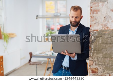 A stylish young entrepreneurial man leaning against a brick wall with a laptop computer in a modern office.