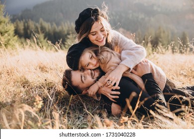 Stylish Young Family In The Autumn Mountains. A Guy And A Girl With Their Daughter Lie And Cuddle On The Grass Against The Background Of The Forest And Mountain Peaks At Sunset.