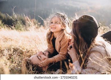 Stylish young family in the autumn mountains. A guy and a girl with their daughter lie and cuddle on the grass against the background of the forest and mountain peaks at sunset.
