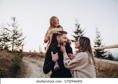 Stylish Young Family In The Autumn Mountains. A Guy And A Girl Together With Their Daughter, Are Standing And Hugging On The Road Against The Background Of A Fence, Forest And Mountain Peaks At Sunset