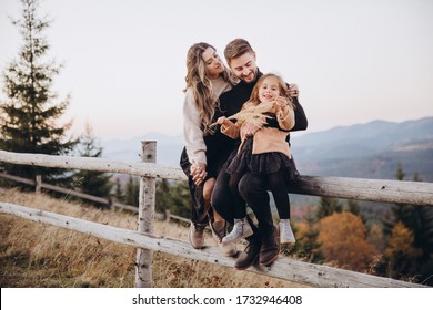 Stylish Young Family In The Autumn Mountains. A Guy And A Girl, Together With Their Daughter, Are Sitting On A Fence Amid A Forest And Mountain Peaks At Sunset.